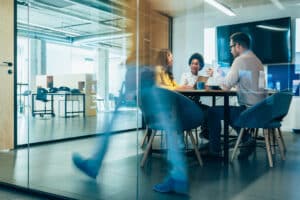 Businesspeople in Conference Room and Colleagues Walking By. Blurred motion of colleagues walking briskly down office hallway as colleagues sit in conference room discussing ideas. Busy modern workplace. Working process in the office, business people working, walking and talking, blurred motion